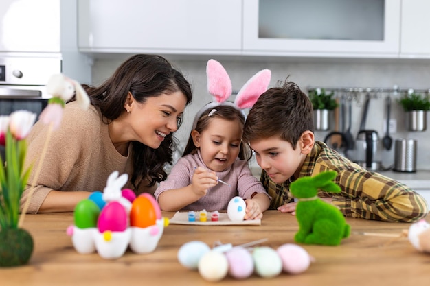 Glückliche Familie malt Ostereier auf dem Küchentisch Mutter, Sohn und Tochter malen Eier Glückliche Familie bereitet sich auf Ostern vor Süßes kleines Mädchen mit Kaninchenohren