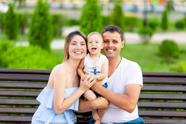 Glückliche Familie Mama und Papa im Park sitzen draußen auf einer Bank im Freien und haben Spaß mit einem Baby im Arm