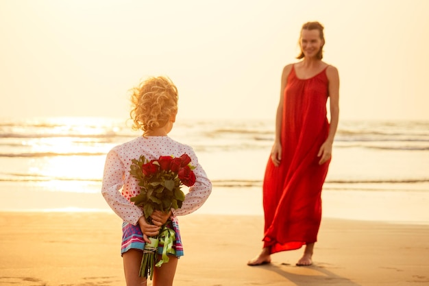 Glückliche Familie mit Blumenstrauß Rosen am Strand am Meer bei Sonnenuntergang Junge schöne erstaunte Mutter und Tochter gratulieren alles Gute zum GeburtstagValentinstag 8. März Internationaler Frauentag