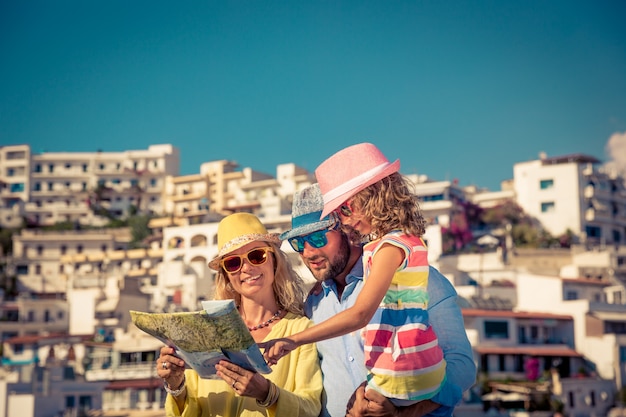 Glückliche Familie mit bunten Sonnenbrillen und Hüten, die eine Karte mit einer europäischen Stadt im Hintergrund halten