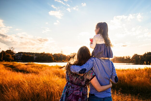 Glückliche Familie mit einer kleinen Tochter in einem Feld in der Natur, nach vorne schauend, Blick von hinten, in den Strahlen des Sonnenuntergangs.