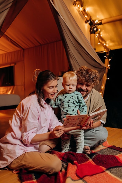 Glückliche Familie mit schönem Baby, das am Sommerabend in der Nähe eines gemütlichen Lagerfeuers zusammen spielt und ein Buch liest Luxus-Campingzelt für Erholung und Erholung im Freien Lifestyle-Konzept