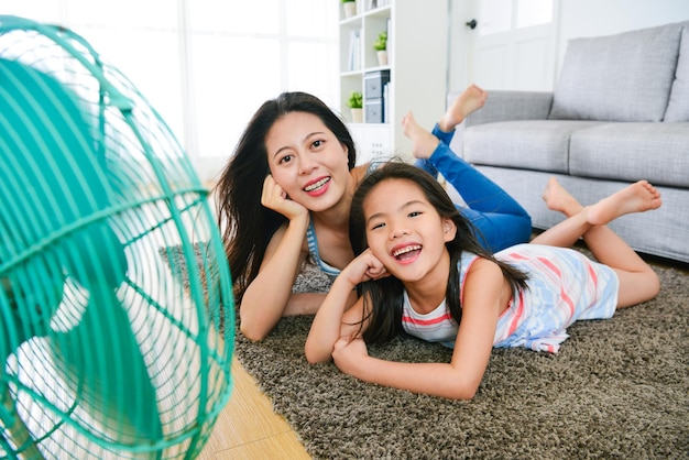 glückliche familie von mutter und tochter, die das gleiche posieren machen und in die kamera schauen, genießen den kühlen wind des elektrischen lüfters im sommertag mit selektivem fokusfoto.