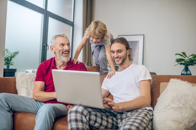 Glückliche Familie. Zwei Männer und ihr Sohn schauen sich etwas auf einem Laptop an und haben Spaß