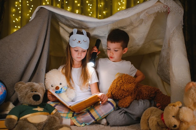 Glückliche Familienkinder, die zu Hause ein Buch mit einer Taschenlampe in einem Zelt lesen