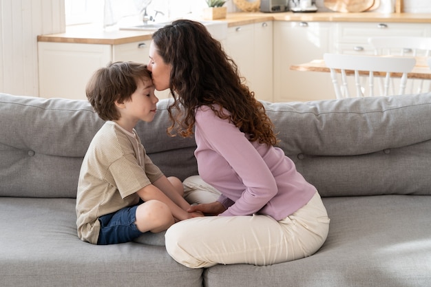 Glückliche familienliebende Mutter, die kleinen Sohn in die Stirn küsst, der mit gekreuzten Beinen zu Hause auf dem Sofa sitzt