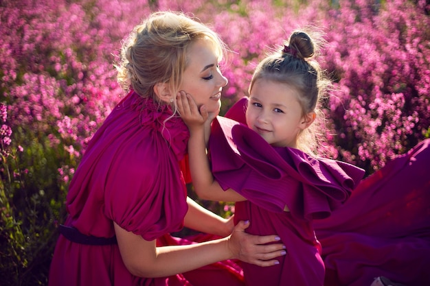 Glückliche Familienmutter und Tochter in lila Kleidern sitzen im Sommer bei Sonnenuntergang auf einem Feld mit Blumen und großen Luftballons