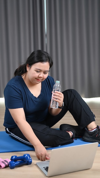 Glückliche fettleibige Frau, die eine Wasserflasche hält und zu Hause Fitnesstrainingsvideos auf dem Laptop ansieht.