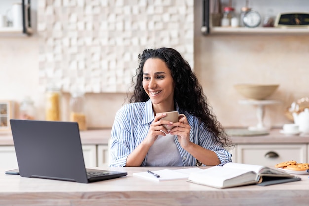 Glückliche Frau des modernen Arbeiter-Heimbüros mit einer Tasse Kaffee trinkend und am Tisch am Laptop arbeitend