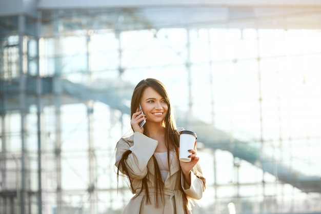 Glückliche Frau, die am Telefon anruft, das nahe Büro steht. Porträt der schönen lächelnden Frau, die Tasse Kaffee in der Hand hält und am Telefon im Freien spricht.