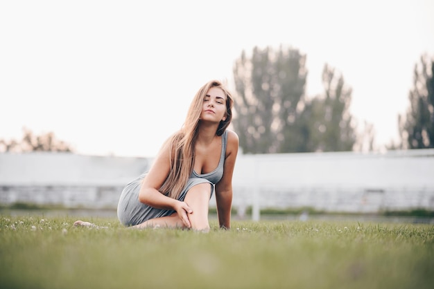 Glückliche Frau, die auf dem Gras sitzt und durchdringend auf die Vorderansicht der Kamera und den Sonnenuntergang im Hintergrund starrt