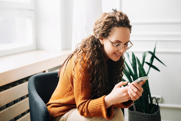 Glückliche Frau, die auf dem Mobiltelefon moderne Apps durchstöbert. Entspannte Frau, die ihr Mobiltelefon zum Online-Shopping nutzt. Junge Frau, die SMS schreibt und auf dem Smartphone im Internet surft. Entspannung zu Hause mit Mobiltelefontechnologie