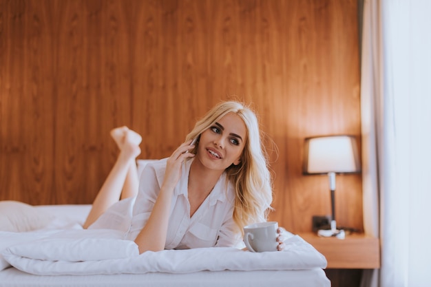 Glückliche Frau, die ein Handy benutzt, während sie auf dem Bett liegt und zu Hause Morgenkaffee trinkt