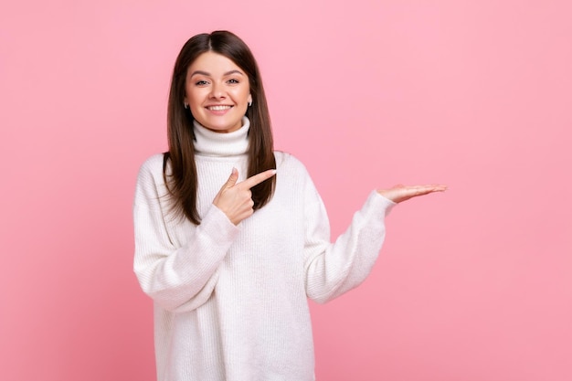 Glückliche Frau, die Kopierraum auf ihrer Handfläche präsentiert, leeren Platz für kommerziellen Text oder Waren zeigt und einen weißen Pullover im lässigen Stil trägt. Studioaufnahme im Innenbereich isoliert auf rosa Hintergrund.