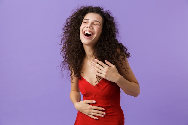 glückliche Frau, die lachendes rotes Kleid trägt und isoliert über violetter Wand steht