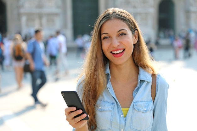 Glückliche Frau, die lächelt und in der Straße mit einem Smartphone geht