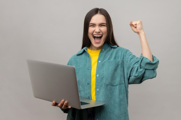 Glückliche Frau, die mit Laptop steht und ihre Faust ballt, um den Erfolg zu feiern und glücklich zu schreien