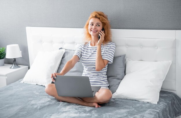 Glückliche Frau, die morgens auf dem Bett mit dem Handy telefoniert Telefonieren, während sie einen Laptop benutzt