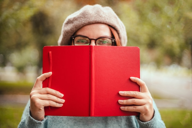 Glückliche Frau, die rote Buchbrille trägt, schaut in die Kamera und lächelt, während sie im Park ruht