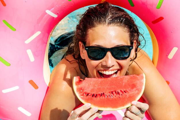 Glückliche Frau, die Sonnenbrille mit roter Wassermelone trägt, genießen im Schwimmbad mit rosa Gummiring