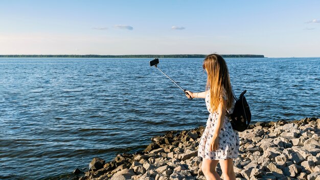 Glückliche Frau, die Videoanruf mit Smartphone und Einbeinstativ am Seehorizont bei Sonnenunterganghintergrund hat