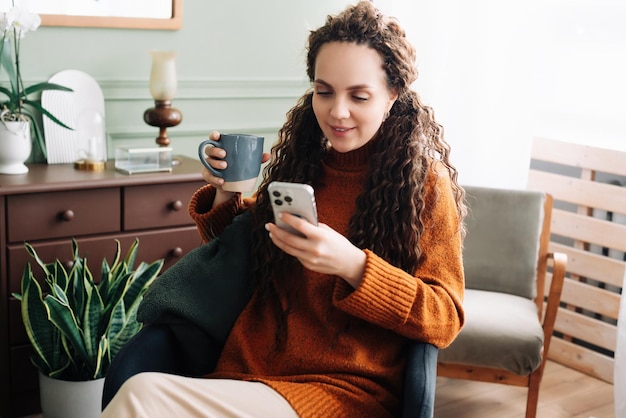 Glückliche Frau, die zu Hause ihr Smartphone benutzt und Kaffee trinkt, lächelnde Dame mit Telefon und Kaffeegenuss