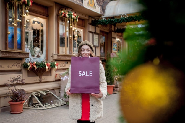 Glückliche Frau hält Papiertüten mit dem Symbol des Verkaufs in den Geschäften mit Weihnachtsverkäufen in der ganzen Stadt. Konzept für Einkaufsferien, Glück, Weihnachtsverkäufe