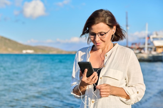 Glückliche Frau im Alter von 40 Jahren in Kopfhörern mit Smartphone am Strand