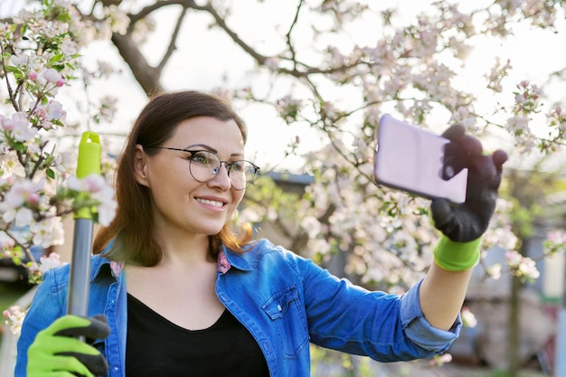 Glückliche Frau im blühenden Garten, die Selfie-Foto auf dem Smartphone macht. Reife Frau mit Gartengeräten in der Hand, Frühlingsgartenarbeit im Hinterhof