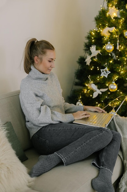 Glückliche Frau im grauen Pullover, die Freunde mit Weihnachten im Videochat auf dem Laptop grüßt. Junge Frau sitzt auf dem Sofa in der Nähe des geschmückten Weihnachtsbaums