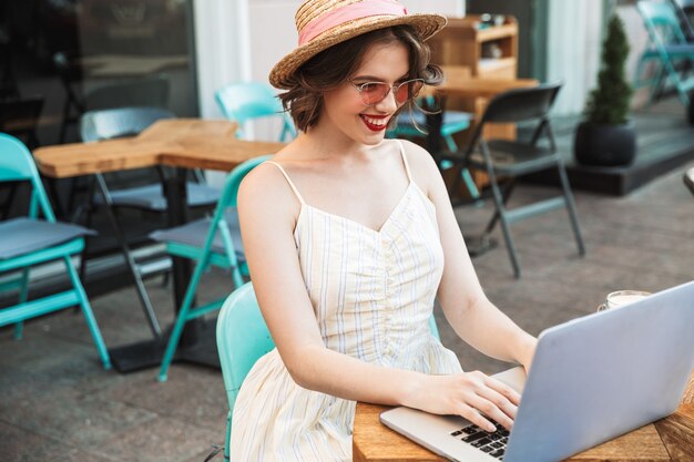 Glückliche Frau im Kleid und im Strohhut unter Verwendung des Laptops