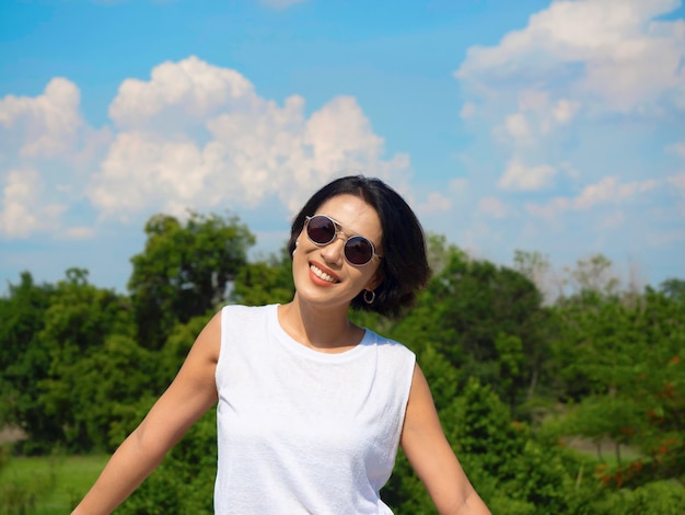 Glückliche Frau im Sommer. Lächelnde schöne asiatische Frau mit kurzen Haaren mit Sonnenbrille und lässigem weißem ärmellosem Hemd, die an einem sonnigen Tag im Sommer auf blauem Himmel, Wolken und grünem Baumhintergrund steht.