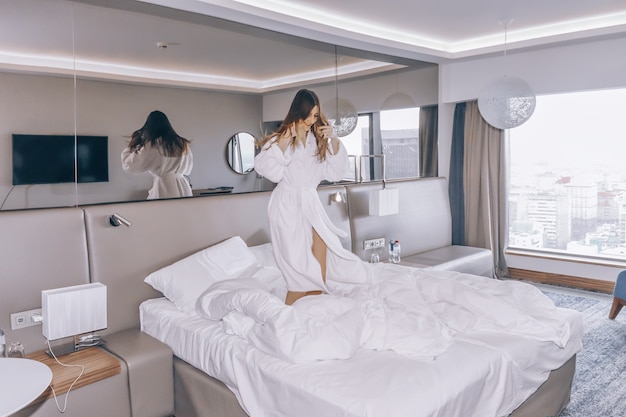 Glückliche Frau im weißen Bademantel springt auf das Bett im Hotelzimmer.