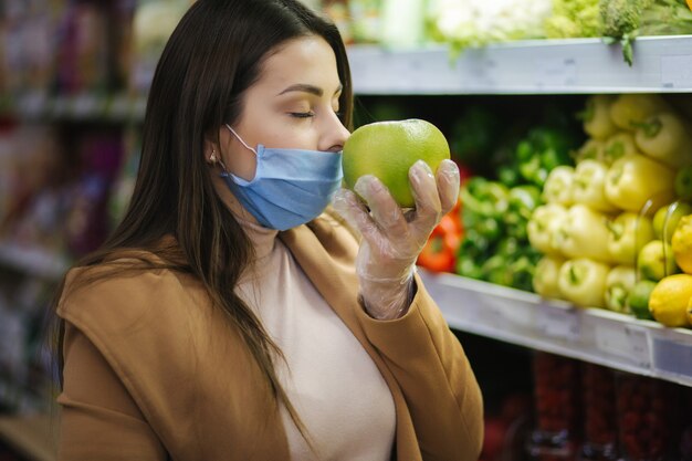 Glückliche Frau in der Schutzmaske, die frisches Gemüse nimmt, während sie durch Lebensmittel im Supermarkt steht