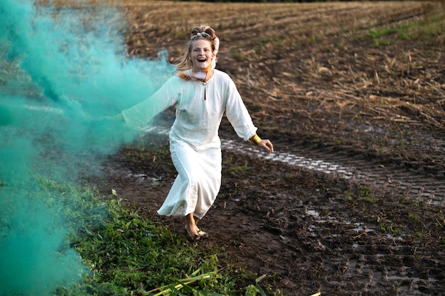 Glückliche Frau in weißem Kleid mit grüner Rauchbombe tanzt auf einem schlammigen Feld Freiheit und Emotionen