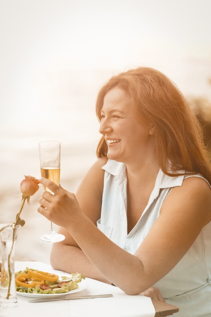 Glückliche Frau lächelt, ein Glas Wein oder Champagner vor dem Hintergrund des Sandstrandes erhebend.