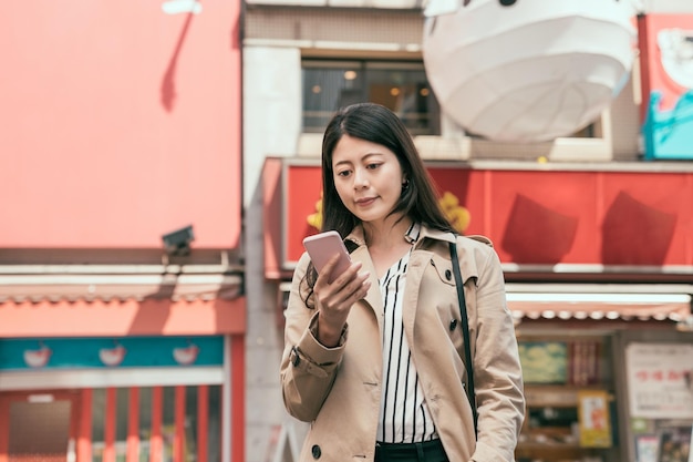 Glückliche Frau lächelt und steht mit Smartphone auf der Straße. junges mädchen in eleganter freizeitkleidung mit handy, das online-karte in dotonbori sucht. Dame, die berühmtes Restaurant online auf dem Handy findet