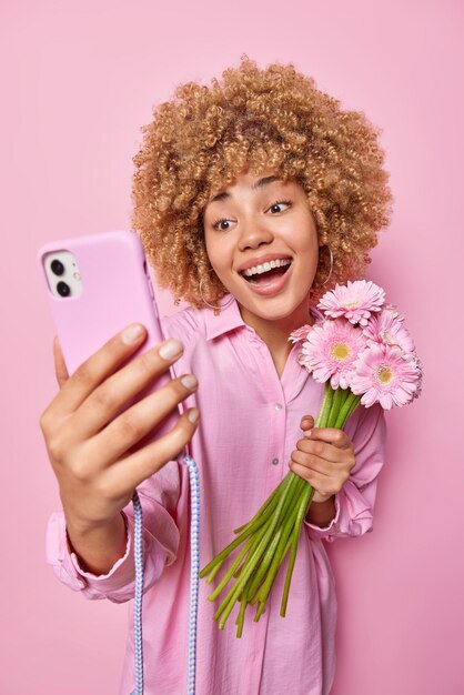 Foto glückliche frau lächelt zahnig macht selfie über smartphone hält gerbera-posen mit lieblingsblumen macht videoanruf trägt hemd isoliert auf rosa wand optimistisches lockiges weibliches modell mit blumenstrauß