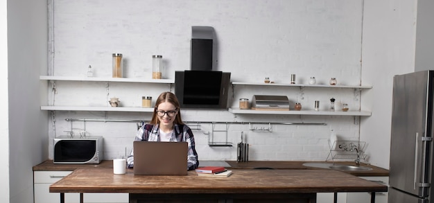 Glückliche frau mit brille, die am morgen zu hause arbeitet. mädchen, das kaffee trinkt. sie benutzt ihren laptop