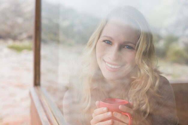 Glückliche Frau mit der Kaffeetasse, die durch Fenster schaut