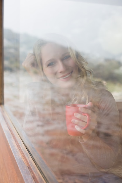 Glückliche Frau mit der Kaffeetasse, die durch Fenster schaut