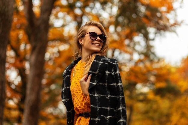 Glückliche Frau mit einem süßen Lächeln mit Zähnen in modischer Herbstkleidung mit einem Strickpullover und einem Mantel in der Natur auf dem Hintergrund eines Baumes mit gelbem Herbstlaub. Lächelndes weibliches hübsches Gesicht