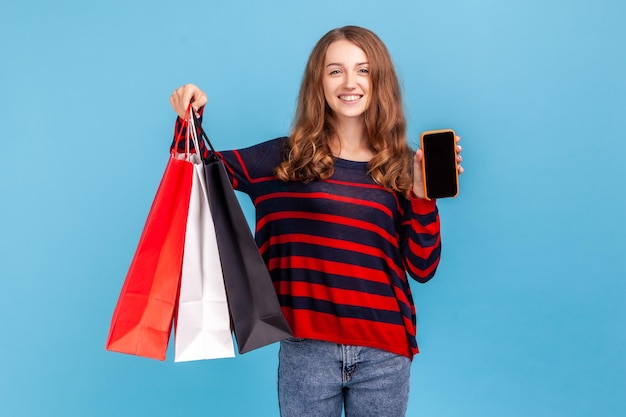 Glückliche Frau mit gestreiftem Pullover im lässigen Stil, die mit zufriedenem Blick steht und Einkaufstaschen und Smartphone mit leerem Bildschirm für Werbung zeigt. Studioaufnahme im Innenbereich isoliert auf blauem Hintergrund.