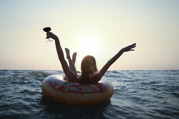 Glückliche Frau mit Glas Wein und aufblasbarem Ring im Meer bei Sonnenuntergang Rückansicht