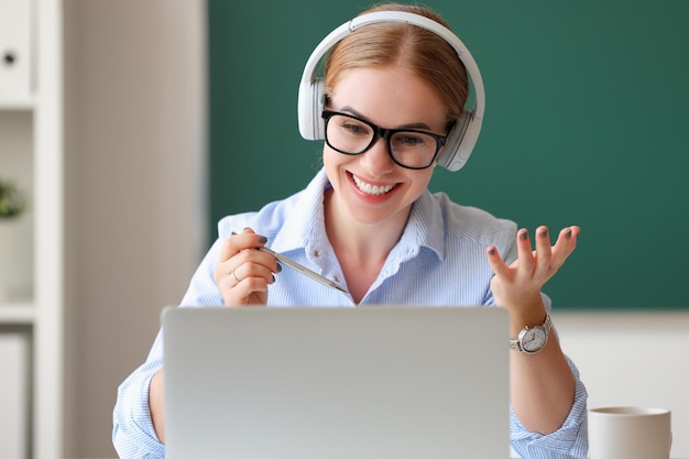 Glückliche Frau mit Kopfhörern und Brille lächelt und spricht mit Schülern, während sie im Klassenzimmer einen Videoanruf tätigt