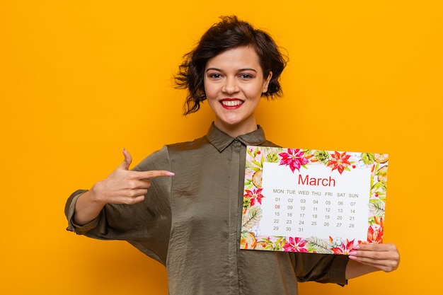 Glückliche Frau mit kurzen Haaren, die den Papierkalender des Monats März hält und mit dem Zeigefinger darauf zeigt, lächelt fröhlich und feiert den internationalen Frauentag 8. März, der über orangefarbenem Hintergrund steht