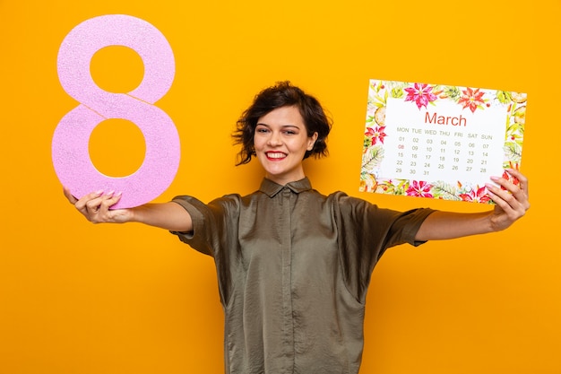 Glückliche Frau mit kurzen Haaren, die den Papierkalender des Monats März und die Nummer acht hält und die Kamera anschaut und fröhlich den internationalen Frauentag am 8. März feiert, der über orangefarbenem Hintergrund steht