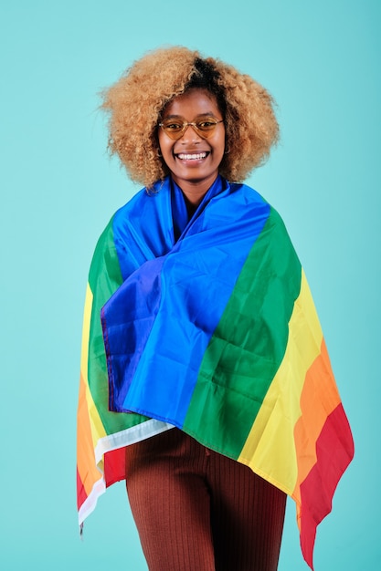 Glückliche Frau mit lockigem Haar, die lächelt, während sie eine Regenbogenflagge der LGBT-Bewegung über einem isolierten Hintergrund hält.