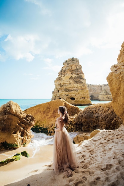 glückliche Frau mit Rücken in einem gelben Kleid, die bei Sonnenuntergang am Strand und am Meer steht