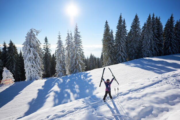 Glückliche Frau mit Ski, der in der Mitte des schneebedeckten Berghangs steht. Sonniger Tag in den Winterferien. Gesamtansicht.